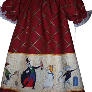May include: A red plaid dress with gold and green accents, featuring a white panel with illustrations of characters from a storybook. The dress has short sleeves with lace trim and a round neckline. The dress is designed for children.