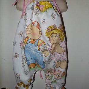 May include: A white sleeveless romper featuring a colorful cartoon print of a boy in blue overalls and a girl in a pink dress surrounded by flowers. The garment has pink tie-up shoulder straps and gathered elastic leg openings with ruffled edges.