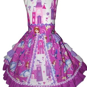 May include: A sleeveless, purple and white dress featuring a princess and castle design. The dress has a ruffled skirt, white lace trim, and a square neckline. The bodice and skirt are adorned with illustrations of castles, princesses, and floral accents.