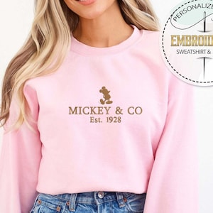 Puede incluir: Una sudadera rosa con una silueta de Mickey Mouse marr&oacute;n y el texto "Mickey & Co. Est. 1928" en la parte delantera.