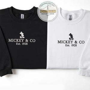 Puede incluir: Dos sudaderas, una negra y otra blanca, con una silueta de Mickey Mouse y el texto "MICKEY & CO Est. 1928" impreso en la parte delantera.