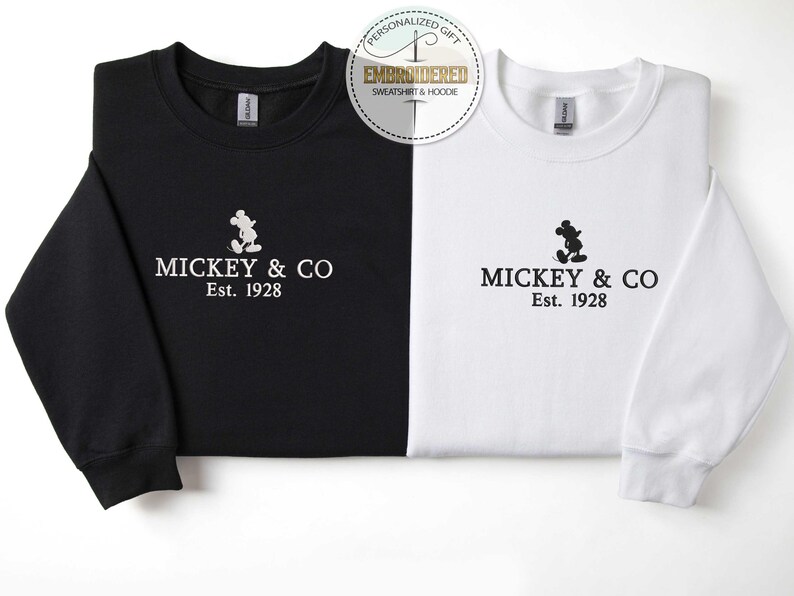 Puede incluir: Dos sudaderas, una negra y otra blanca, con una silueta de Mickey Mouse y el texto "MICKEY & CO Est. 1928" impreso en la parte delantera.