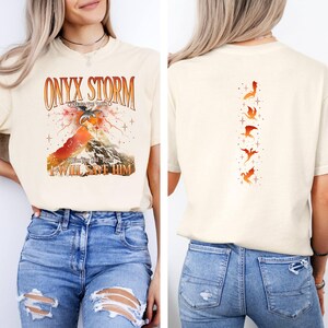 May include: A cream colored t-shirt with the text "ONYX STORM" and a graphic of a mountain with a dragon flying over it. The back of the shirt has a vertical row of five orange dragons.
