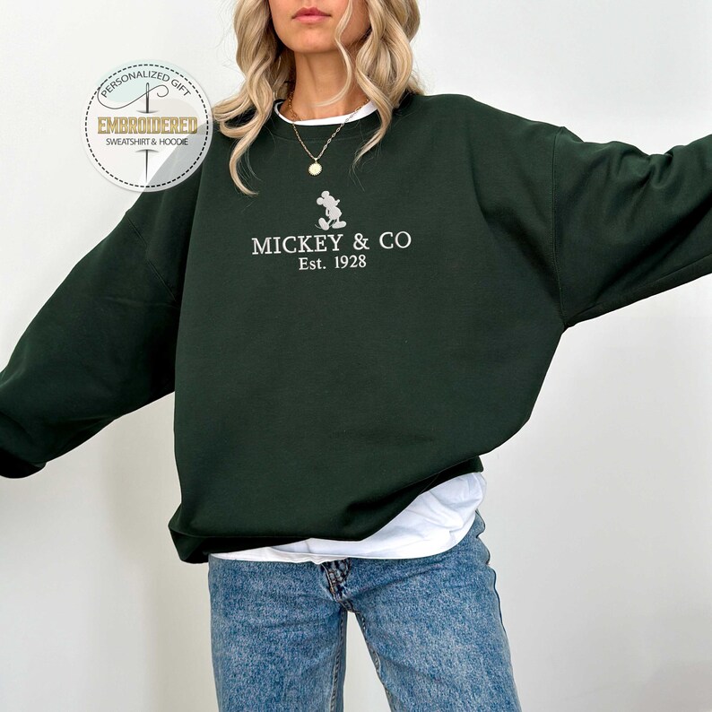 Puede incluir: Una sudadera verde oscuro con un Mickey Mouse bordado en blanco y el texto "MICKEY & CO Est. 1928".