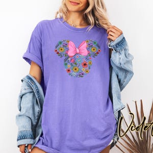 Puede incluir: Una camiseta de color violeta con un dise&ntilde;o floral de Minnie Mouse y un lazo rosa. El dise&ntilde;o est&aacute; formado por varias flores coloridas. La palabra "Violet" est&aacute; escrita en cursiva en la parte inferior derecha de la imagen.