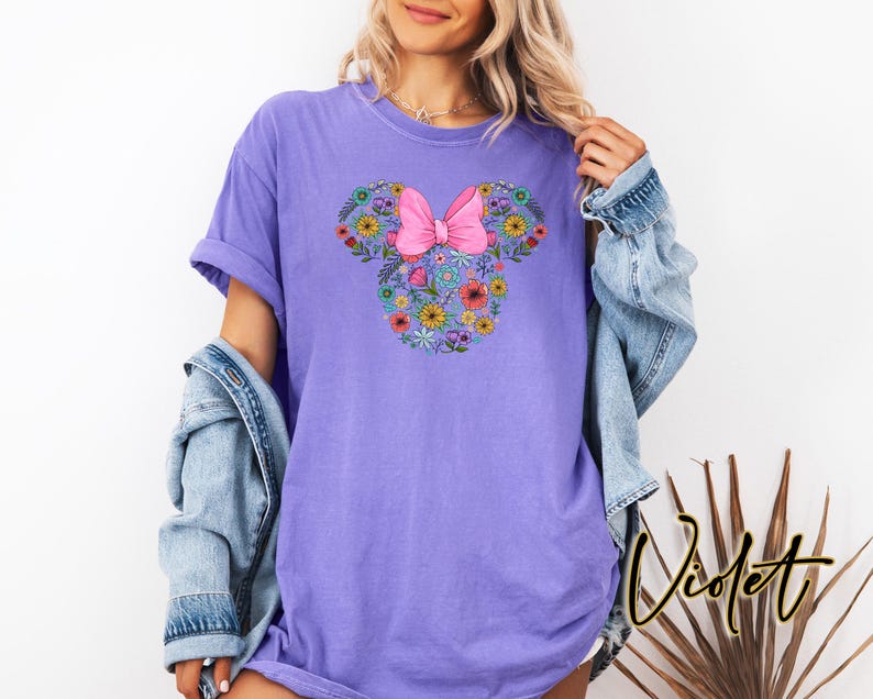 Puede incluir: Una camiseta de color violeta con un dise&ntilde;o floral de Minnie Mouse y un lazo rosa. El dise&ntilde;o est&aacute; formado por varias flores coloridas. La palabra "Violet" est&aacute; escrita en cursiva en la parte inferior derecha de la imagen.