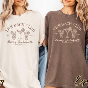 May include: Two t-shirts with a brown and white design. The text on the shirts reads "The Bach Club Est. 2024 Karen's Bachelorette San Diego, CA". One shirt is ivory and the other is espresso.