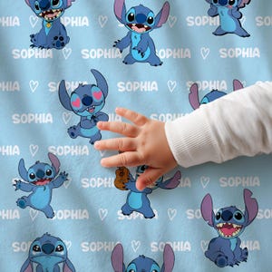 Op de afbeelding: Lichtblauwe deken met meerdere cartoonafbeeldingen van Stitch, met de naam "SOPHIA" en harten. Een babyhand is zichtbaar, die de deken aanraakt. De deken is zacht en waarschijnlijk van fleece.