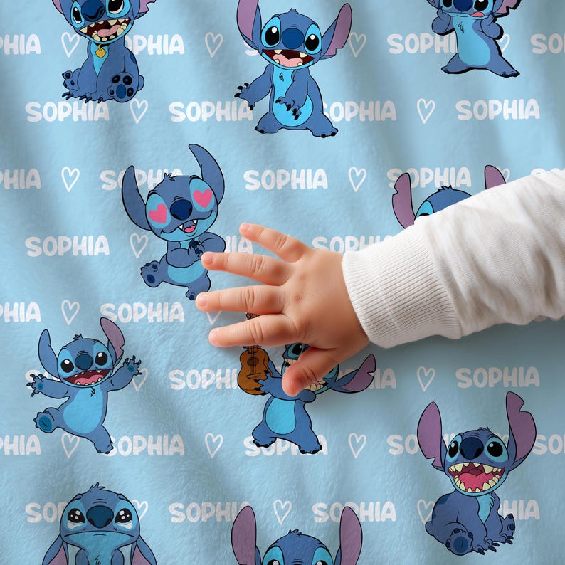 Op de afbeelding: Lichtblauwe deken met meerdere cartoonafbeeldingen van Stitch, met de naam "SOPHIA" en harten. Een babyhand is zichtbaar, die de deken aanraakt. De deken is zacht en waarschijnlijk van fleece.