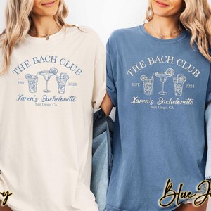 May include: Two t-shirts with the text "The Bach Club Est 2024 Karen's Bachelorette San Diego, CA" and a graphic of three cocktail glasses. One shirt is ivory and the other is blue jean.
