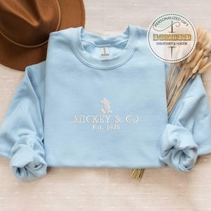 Puede incluir: Una sudadera azul claro con un dise&ntilde;o de Mickey Mouse bordado en blanco y el texto "Mickey & Co. Est. 1928".