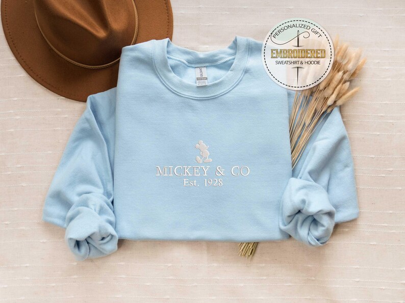 Puede incluir: Una sudadera azul claro con un dise&ntilde;o de Mickey Mouse bordado en blanco y el texto "Mickey & Co. Est. 1928".