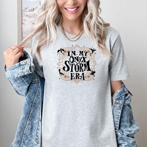 Puede incluir: Una camiseta gris con un dise&ntilde;o gr&aacute;fico que dice "In My Onyx Storm Era" con un marco dorado y dragones negros.