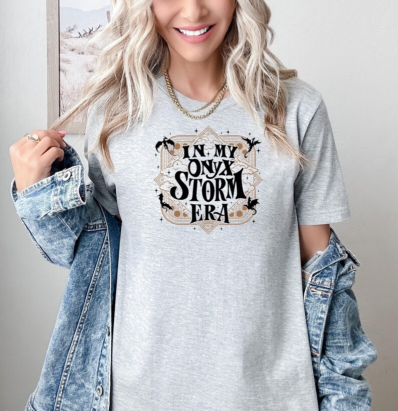 Puede incluir: Una camiseta gris con un dise&ntilde;o gr&aacute;fico que dice "In My Onyx Storm Era" con un marco dorado y dragones negros.
