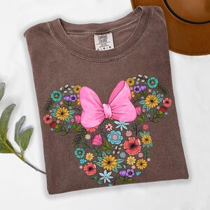 Puede incluir: Camiseta Comfort Colors marr&oacute;n con un dise&ntilde;o floral de Minnie Mouse. El dise&ntilde;o incluye un gran lazo rosa y flores coloridas en tonos amarillos, azules, morados y rojos. La camiseta es talla L-G-G.