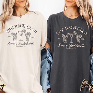 May include: Two women's t-shirts with a black graphic design that says "The Bach Club" with a date and the text "Karen's Bachelorette" with a city and state. The t-shirts are in ivory and pepper colors.