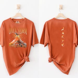 May include: A burnt orange t-shirt with a graphic print of a mountain and the text "ONYX STORM - Brave The Dark - I Will Not Let You Down - I Will Save Him". The back of the shirt has a graphic print of five flying birds.