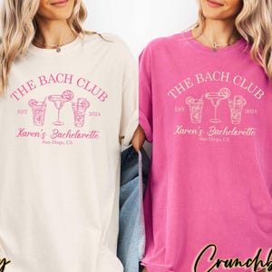 May include: Two white t-shirts with pink text that reads "The Bach Club Est. 2024 Karen's Bachelorette San Diego, CA". The shirts feature a graphic of three cocktail glasses with pink outlines.