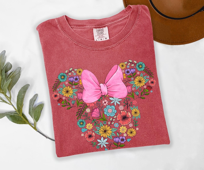 Puede incluir: Una camiseta roja Comfort Colors con un dise&ntilde;o floral de Minnie Mouse. El dise&ntilde;o incluye un lazo rosa y un arreglo floral de varios colores, incluyendo amarillo, azul y morado. La camiseta est&aacute; doblada y se muestra sobre una superficie blanca.