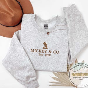 Puede incluir: Una sudadera gris con una silueta de Mickey Mouse bordada en marr&oacute;n y el texto "MICKEY & CO Est. 1928".