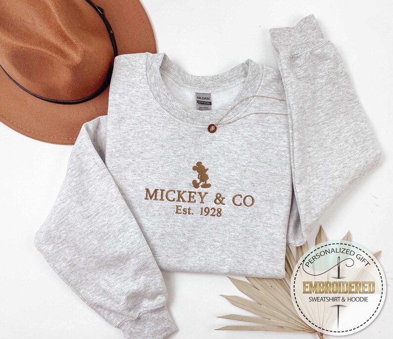Puede incluir: Una sudadera gris con una silueta de Mickey Mouse bordada en marr&oacute;n y el texto "MICKEY & CO Est. 1928".