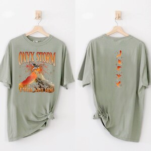 May include: A sage green t-shirt with a graphic print of a dragon and a mountain. The text "ONYX STORM" is printed in orange and black. The text "I WILL SAVE HIM" is printed in orange. The back of the shirt has a graphic print of five orange birds.