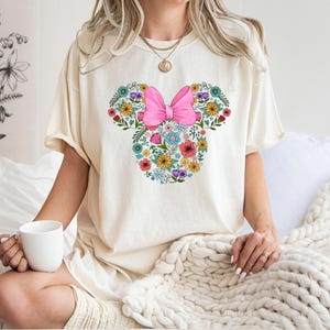 Puede incluir: Camiseta color crema con un dise&ntilde;o floral de Minnie Mouse. El dise&ntilde;o incluye un lazo rosa y una variedad de flores coloridas. La persona sostiene una taza blanca. La camiseta es de estilo c&oacute;modo e informal.