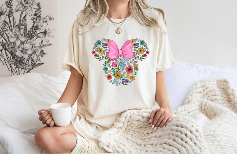Puede incluir: Camiseta color crema con un dise&ntilde;o floral de Minnie Mouse. El dise&ntilde;o incluye un lazo rosa y una variedad de flores coloridas. La persona sostiene una taza blanca. La camiseta es de estilo c&oacute;modo e informal.