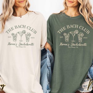 May include: Two women's t-shirts with a green and ivory color scheme. The shirts feature a graphic design of a cocktail glass with the text "The Bach Club Est. 2024 Karen's Bachelorette San Diego, CA".