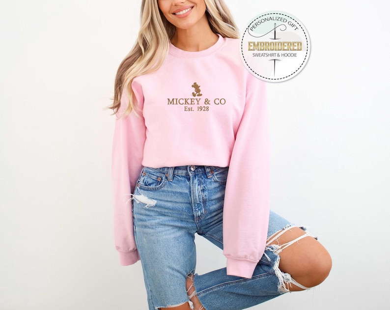Puede incluir: Una sudadera rosa con una silueta de Mickey Mouse marr&oacute;n y el texto "Mickey & Co. Est. 1928" en la parte delantera.