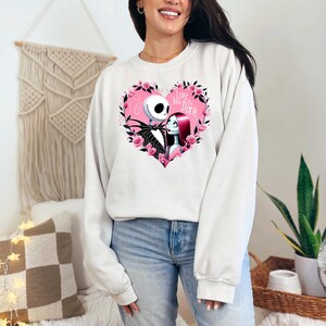 Może przedstawiać: Biała bluza z nadrukiem r&oacute;żowego serca przedstawiającego Jacka Skellingtona i Sally z filmu Koszmar przed świętami. Tekst w sercu brzmi "Love You To Death."