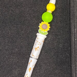 May include: A white pen with a gold leaf design and a sunflower charm. The pen has green and yellow beads at the top.