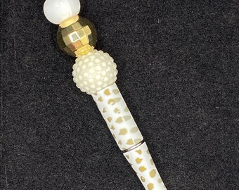 May include: A decorative pen with a white and gold spotted barrel. The pen's top features a series of beads in white, gold, and cream colors. The pen has a silver tip and is set against a black background.
