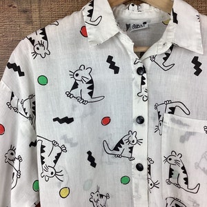 K&ouml;nnte beinhalten: Wei&szlig;es Button-Down-Hemd mit einem verspielten Katzenmuster. Das Hemd zeigt schwarze Katzenillustrationen mit gestreiften K&ouml;rpern, zusammen mit roten, gr&uuml;nen und gelben Kreisen und schwarzen Zickzackformen. Das Hemd hat schwarze Kn&ouml;pfe und eine Tasche.