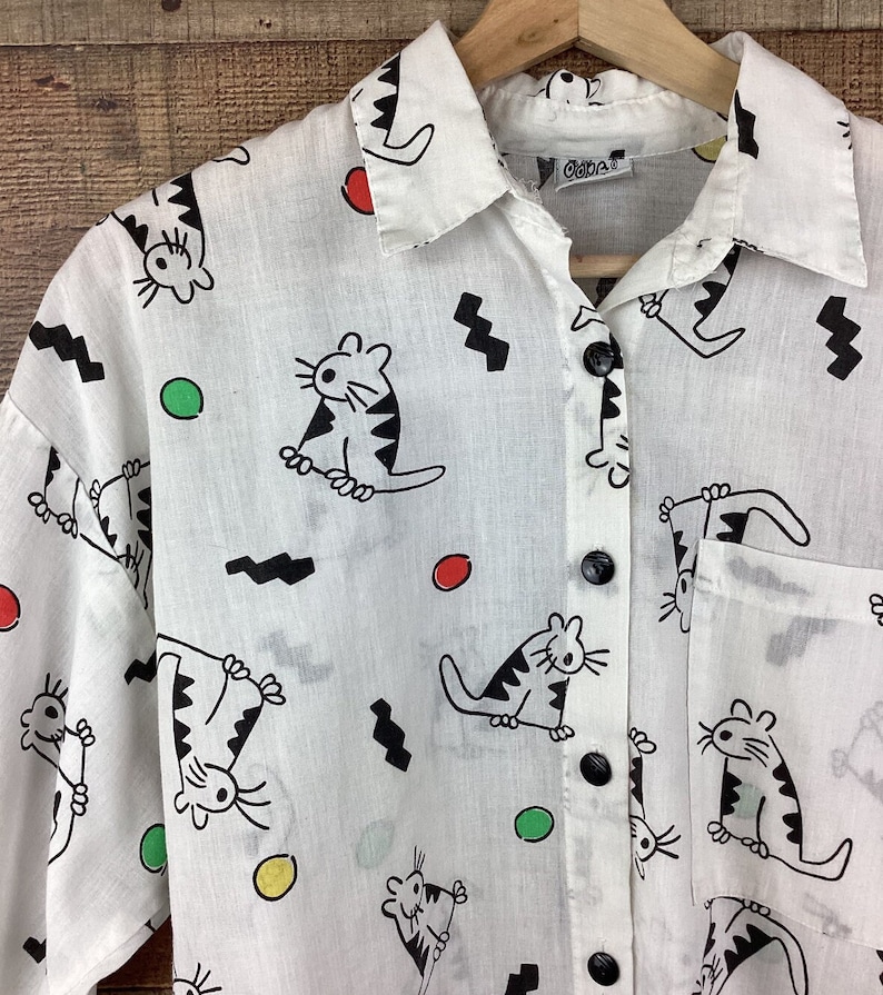 K&ouml;nnte beinhalten: Wei&szlig;es Button-Down-Hemd mit einem verspielten Katzenmuster. Das Hemd zeigt schwarze Katzenillustrationen mit gestreiften K&ouml;rpern, zusammen mit roten, gr&uuml;nen und gelben Kreisen und schwarzen Zickzackformen. Das Hemd hat schwarze Kn&ouml;pfe und eine Tasche.