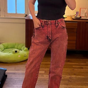 May include: A person wearing a black t-shirt and red-dyed jeans. The jeans have a classic five-pocket design. The person is also wearing black and gray checkered slip-on sneakers.