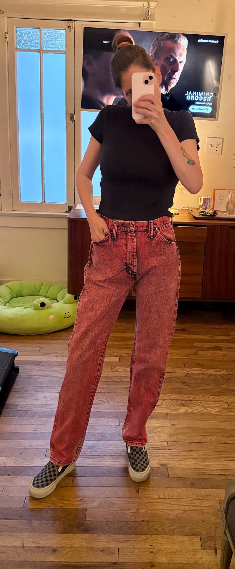 May include: A person wearing a black t-shirt and red-dyed jeans. The jeans have a classic five-pocket design. The person is also wearing black and gray checkered slip-on sneakers.