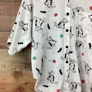 K&ouml;nnte beinhalten: Wei&szlig;es Button-Down-Hemd mit einem verspielten Katzenmuster. Das Hemd zeigt schwarze Katzenillustrationen mit gestreiften K&ouml;rpern sowie schwarzen Zickzacklinien und farbigen Punkten. Es hat eine Brusttasche und schwarze Kn&ouml;pfe.