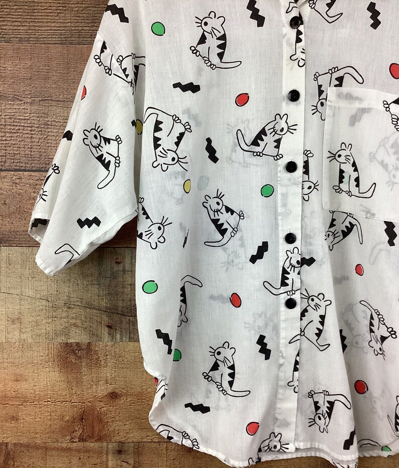 K&ouml;nnte beinhalten: Wei&szlig;es Button-Down-Hemd mit einem verspielten Katzenmuster. Das Hemd zeigt schwarze Katzenillustrationen mit gestreiften K&ouml;rpern sowie schwarzen Zickzacklinien und farbigen Punkten. Es hat eine Brusttasche und schwarze Kn&ouml;pfe.