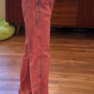 May include: Red acid wash jeans with a black top and checkered slip-on sneakers. The jeans have a classic five-pocket design and a straight leg fit. The sneakers have a black and white checkered pattern.
