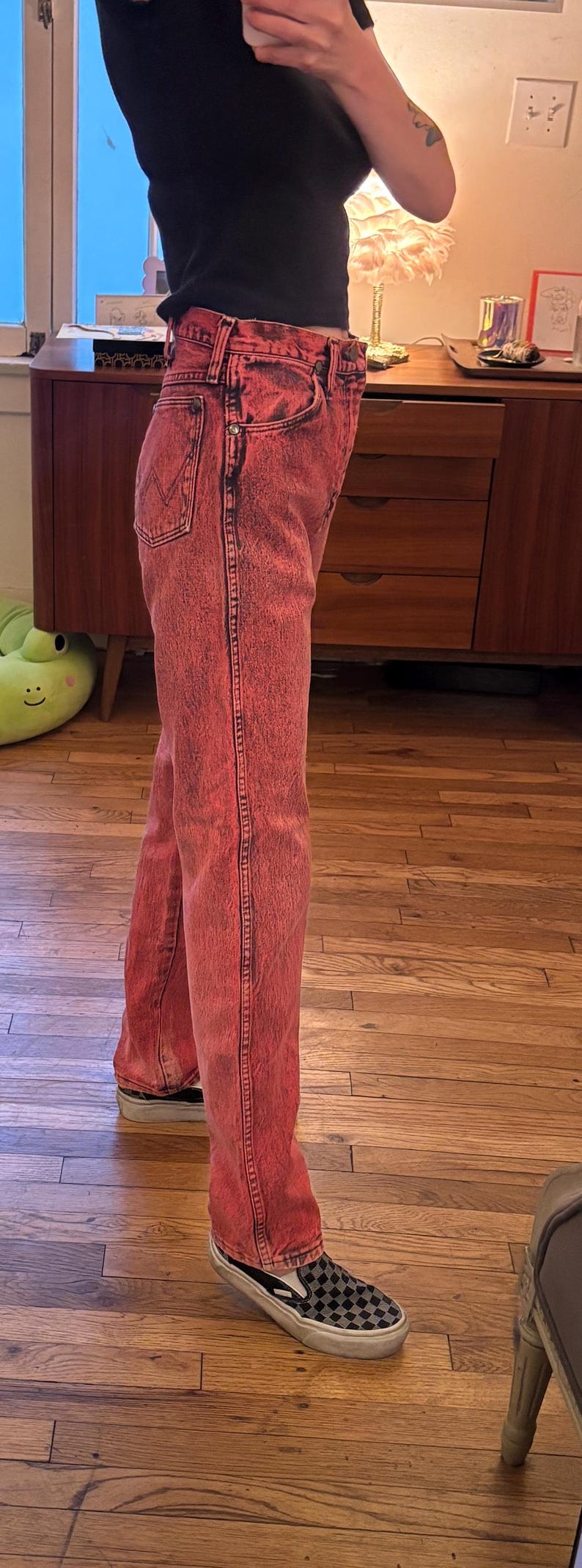 May include: Red acid wash jeans with a black top and checkered slip-on sneakers. The jeans have a classic five-pocket design and a straight leg fit. The sneakers have a black and white checkered pattern.