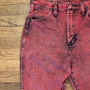 May include: Close-up of acid-wash red denim jeans. The jeans have a classic five-pocket design with a button and zipper closure. The fabric has a faded, textured appearance.