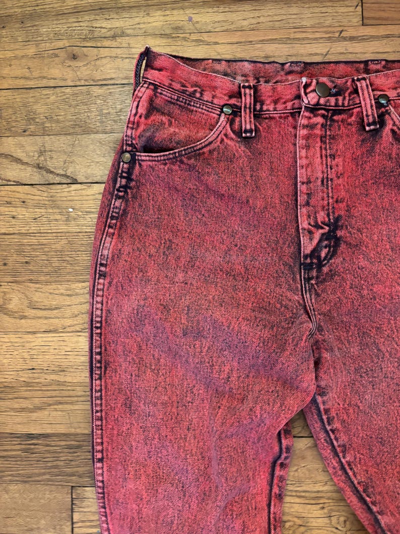 May include: Close-up of acid-wash red denim jeans. The jeans have a classic five-pocket design with a button and zipper closure. The fabric has a faded, textured appearance.