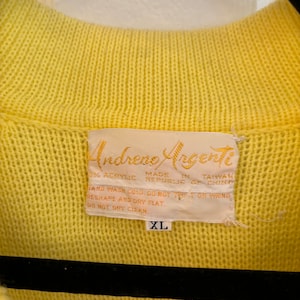 May include: Close-up of a yellow knit sweater with a ribbed collar. The label reads "Andreno Argenti" and includes care instructions and the size "XL". The sweater is made of 33% acrylic and made in Taiwan.