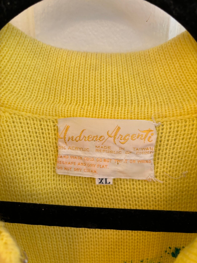 May include: Close-up of a yellow knit sweater with a ribbed collar. The label reads "Andreno Argenti" and includes care instructions and the size "XL". The sweater is made of 33% acrylic and made in Taiwan.