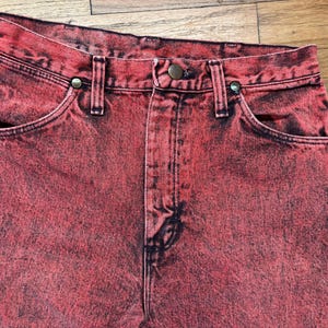 May include: Close-up of red denim jeans with a faded, distressed appearance. The jeans feature classic five-pocket styling, a button and zipper closure, and belt loops. The fabric has a mottled, vintage look.