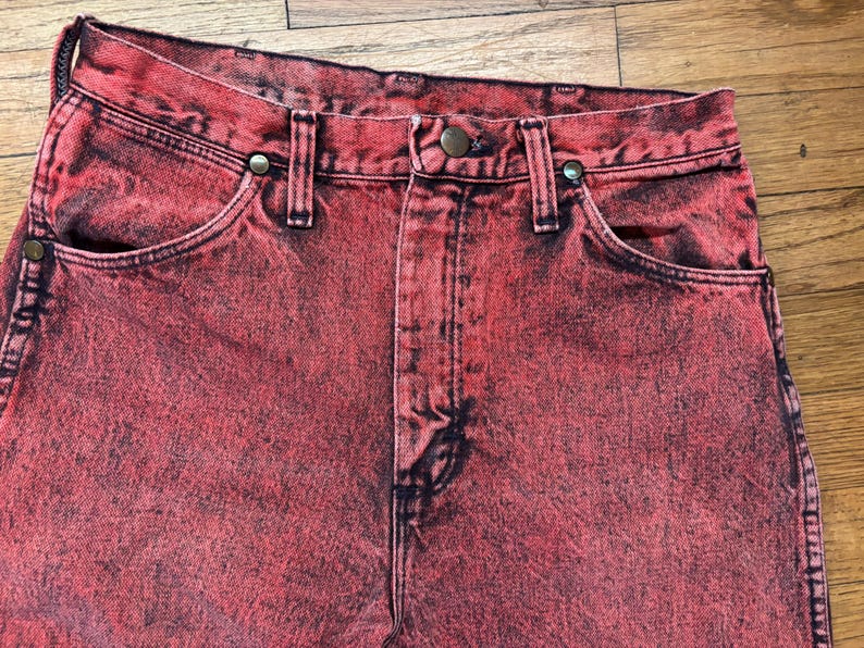 May include: Close-up of red denim jeans with a faded, distressed appearance. The jeans feature classic five-pocket styling, a button and zipper closure, and belt loops. The fabric has a mottled, vintage look.