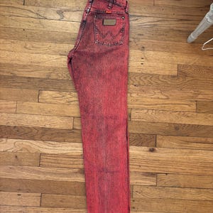 May include: Red acid wash denim jeans with a classic five-pocket design. The jeans feature a brown leather patch on the back pocket and the Wrangler logo. The jeans are laid flat on a wooden surface.