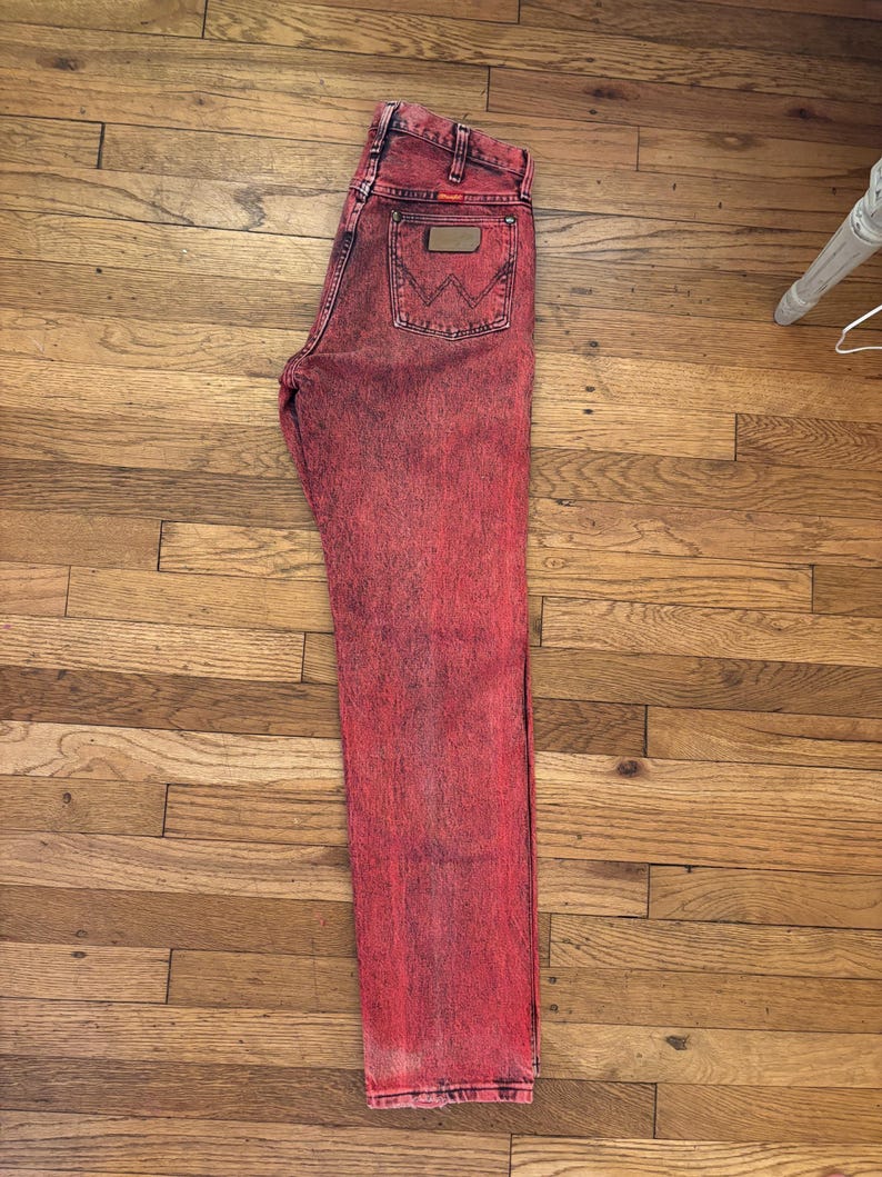 May include: Red acid wash denim jeans with a classic five-pocket design. The jeans feature a brown leather patch on the back pocket and the Wrangler logo. The jeans are laid flat on a wooden surface.