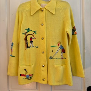 May include: A bright yellow cardigan with a collared neckline and gold buttons. The sweater features embroidered golf-themed designs, including golfers, trees, and golf clubs. It has two front pockets and long sleeves.
